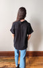 Person wearing a black shirt and blue jeans standing against a white wall on a wooden floor.
