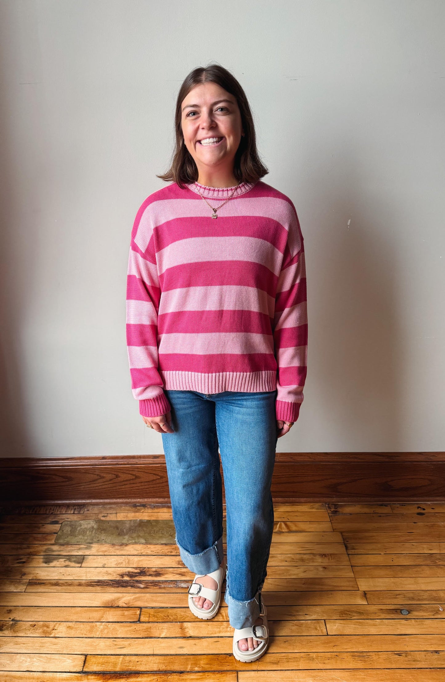 A woman smiling and standing in a room with a light gray wall and wooden floor, wearing a pink and white striped knit sweater, blue jeans, and white slide sandals.