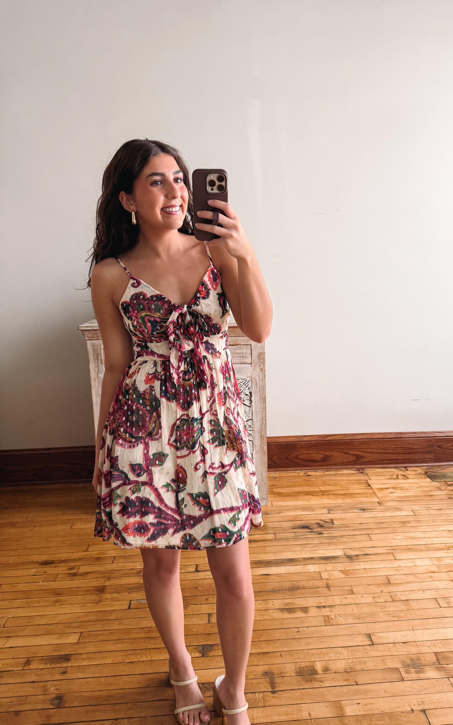 Woman taking a mirror selfie wearing a floral dress in a room with wooden flooring.