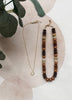A gold necklace with a small circular pendant and a matching beaded necklace featuring brown, dark brown, and cream beads, styled on a light beige textured surface with green leaves.