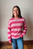 A smiling woman with shoulder-length brown hair stands indoors wearing a pink and light pink striped knit sweater and blue jeans against a plain light wall.