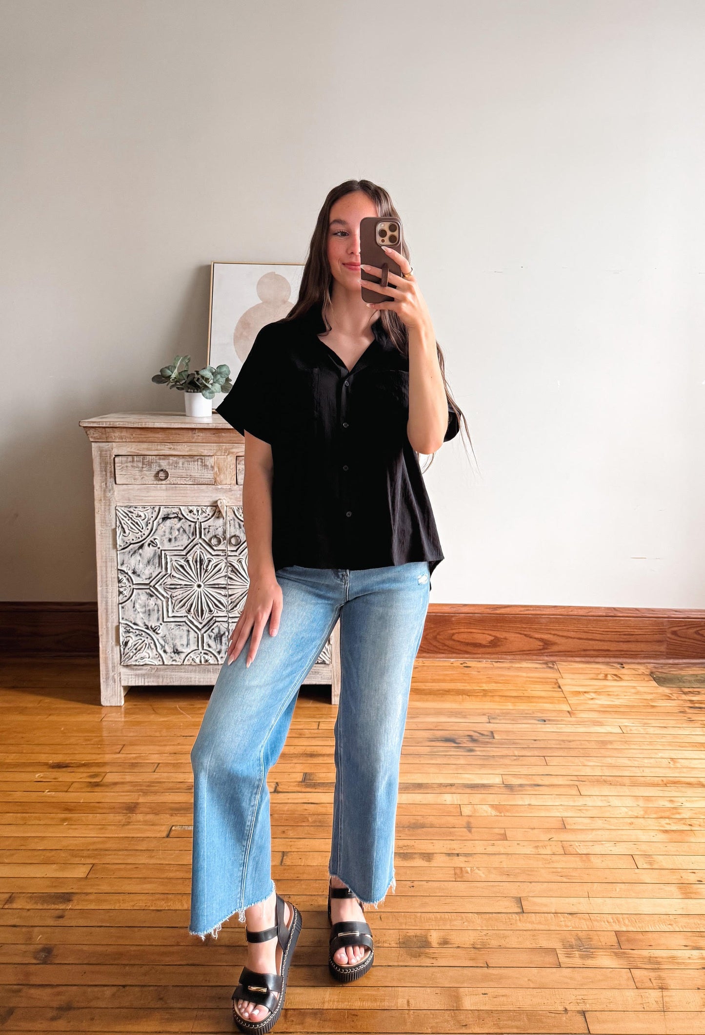 Person taking a mirror selfie wearing a black shirt and blue jeans in a room with wooden flooring and a white cabinet.