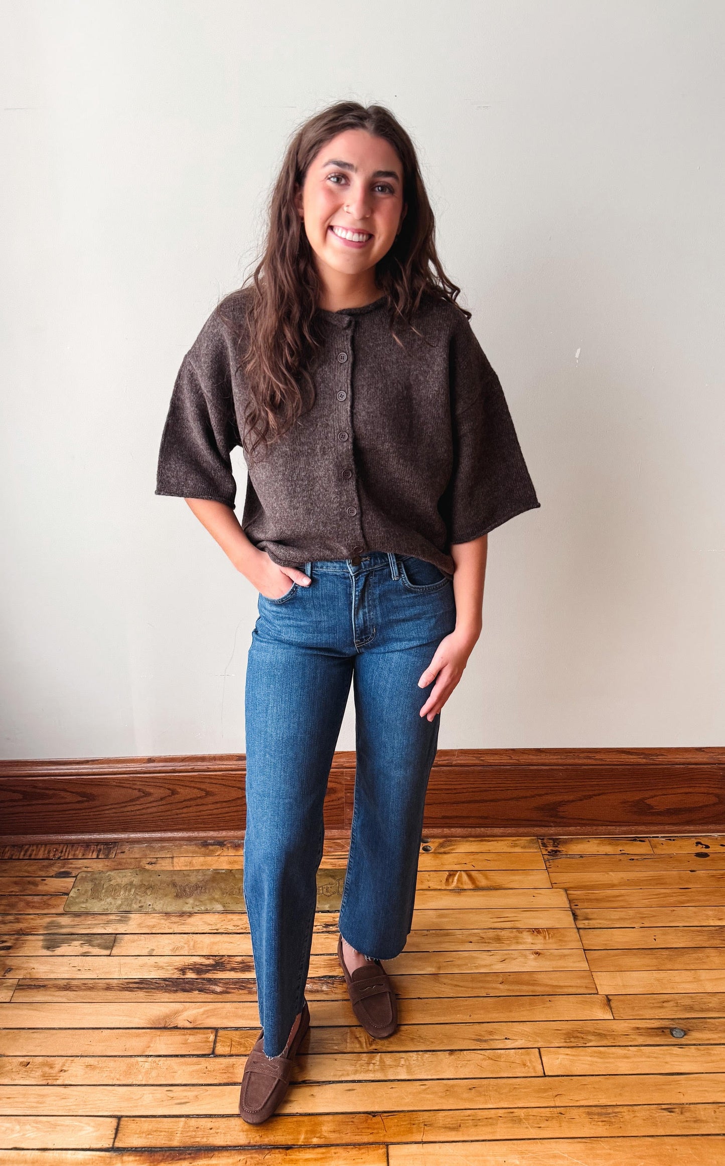 A woman stands in a room with a light beige wall and wooden floor, wearing a dark brown textured short-sleeve button-up top, blue jeans, and brown suede loafers.