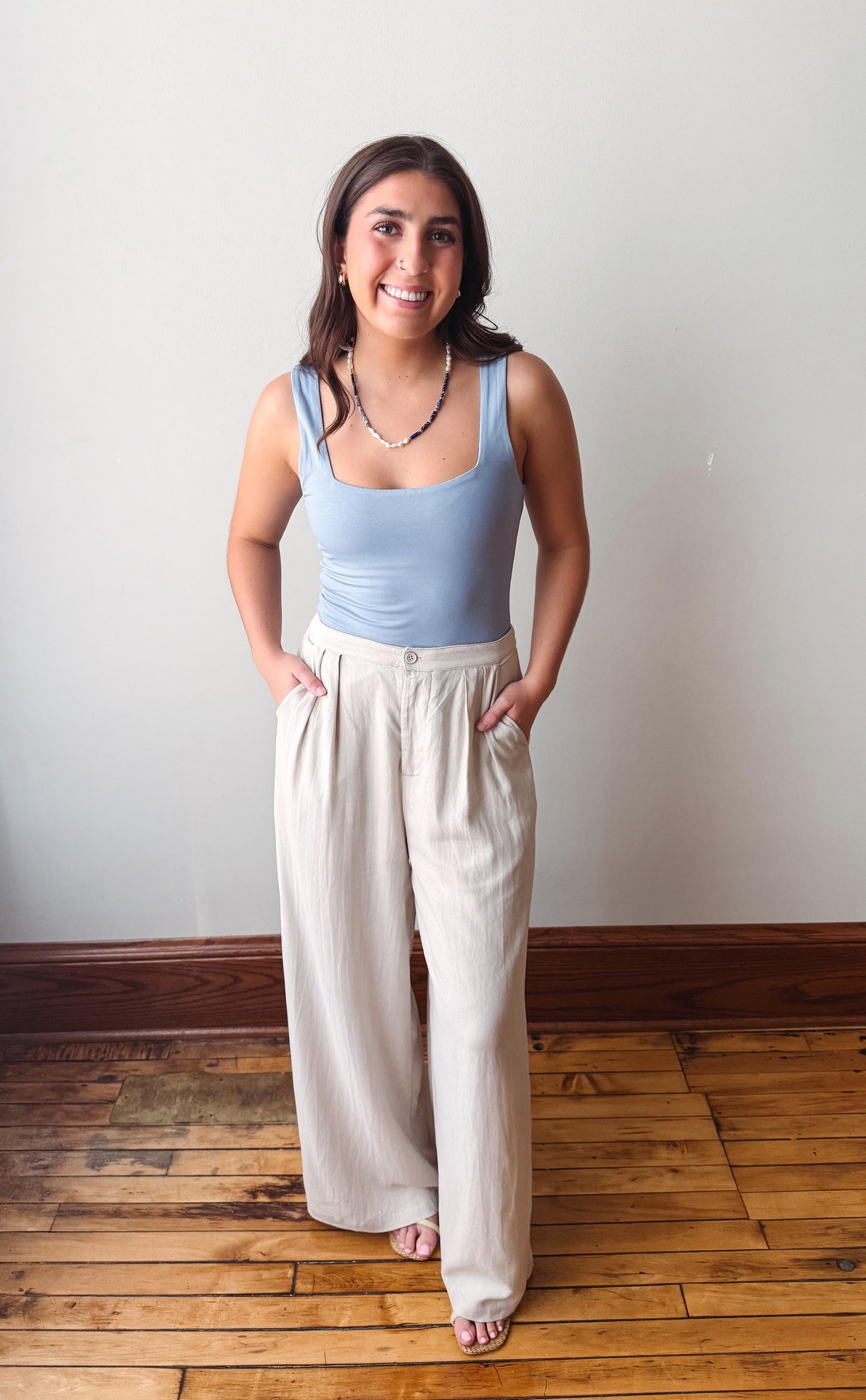 Woman wearing a light blue tank top and beige wide-leg pants standing on a wooden floor.