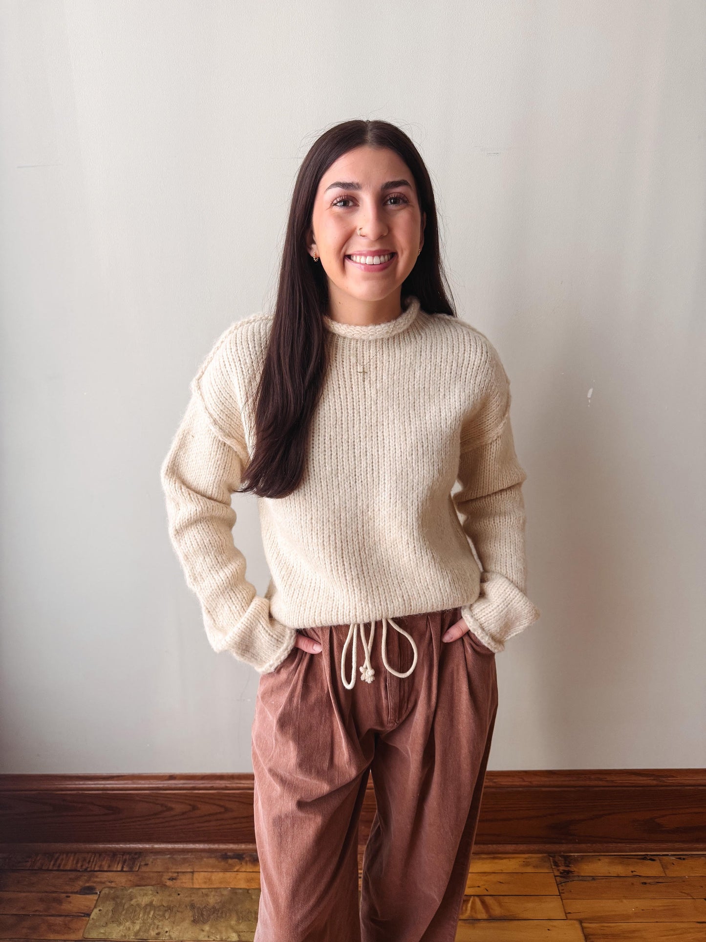 A woman with long dark hair stands indoors wearing a cream ribbed knit sweater and brown drawstring pants against a plain light wall and wood floor.