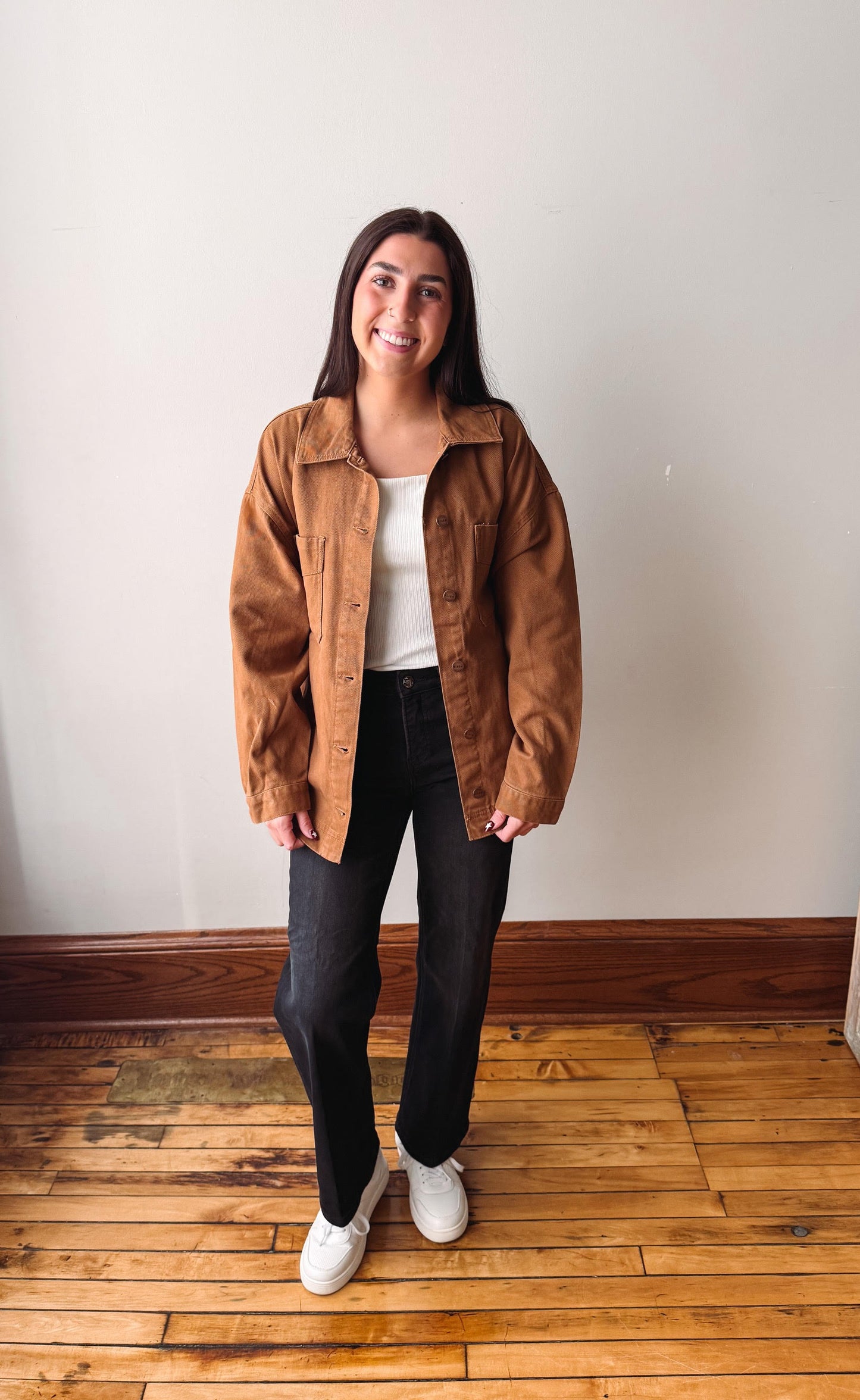 A woman stands in a room with a white wall and wooden floor, wearing a brown corduroy jacket over a white top, black jeans, and white sneakers.