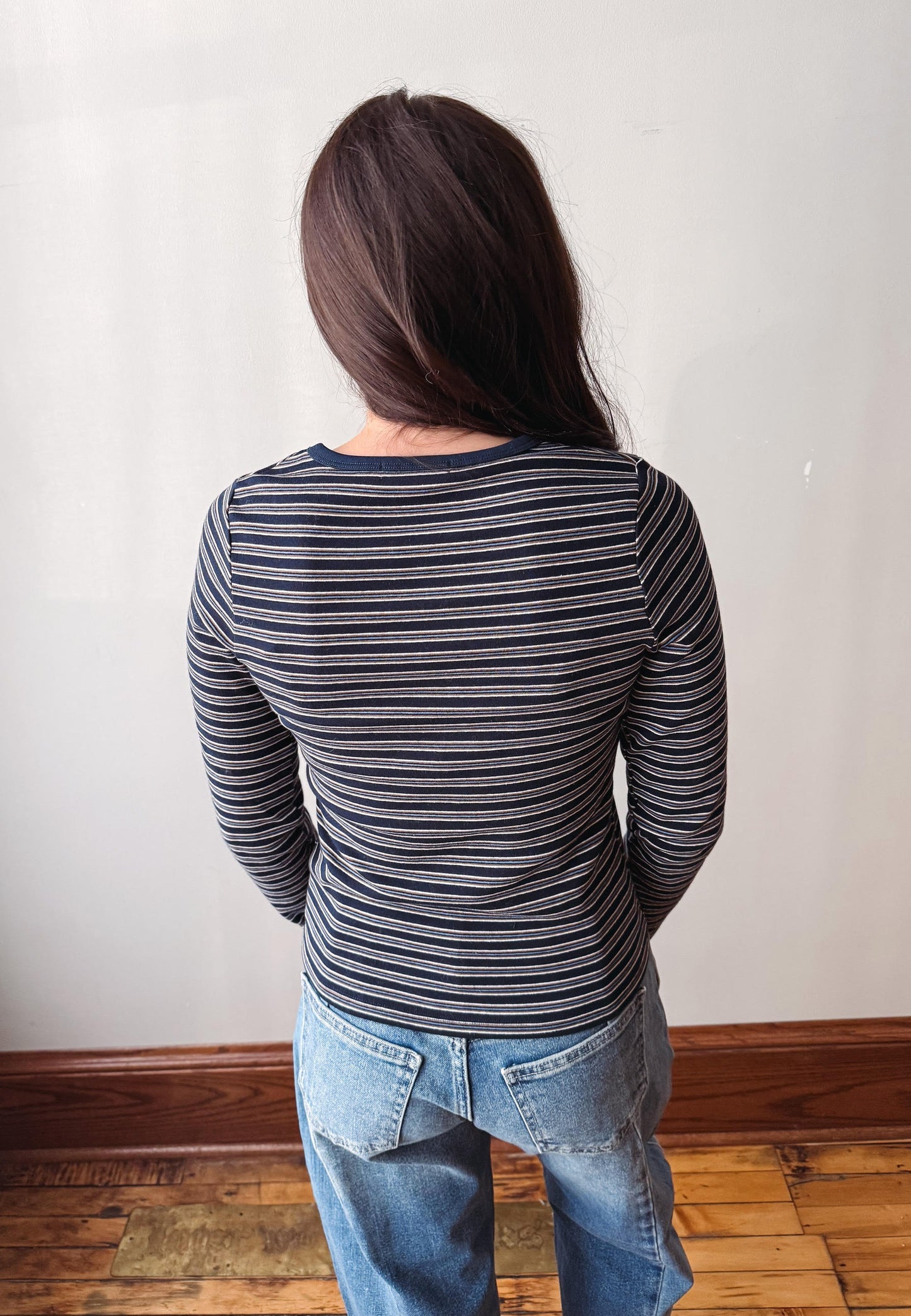A person viewed from behind wearing a navy and gray striped long-sleeve top with blue jeans, standing against an off-white wall on a wooden floor.