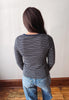 A person viewed from behind wearing a navy and gray striped long-sleeve top with blue jeans, standing against an off-white wall on a wooden floor.