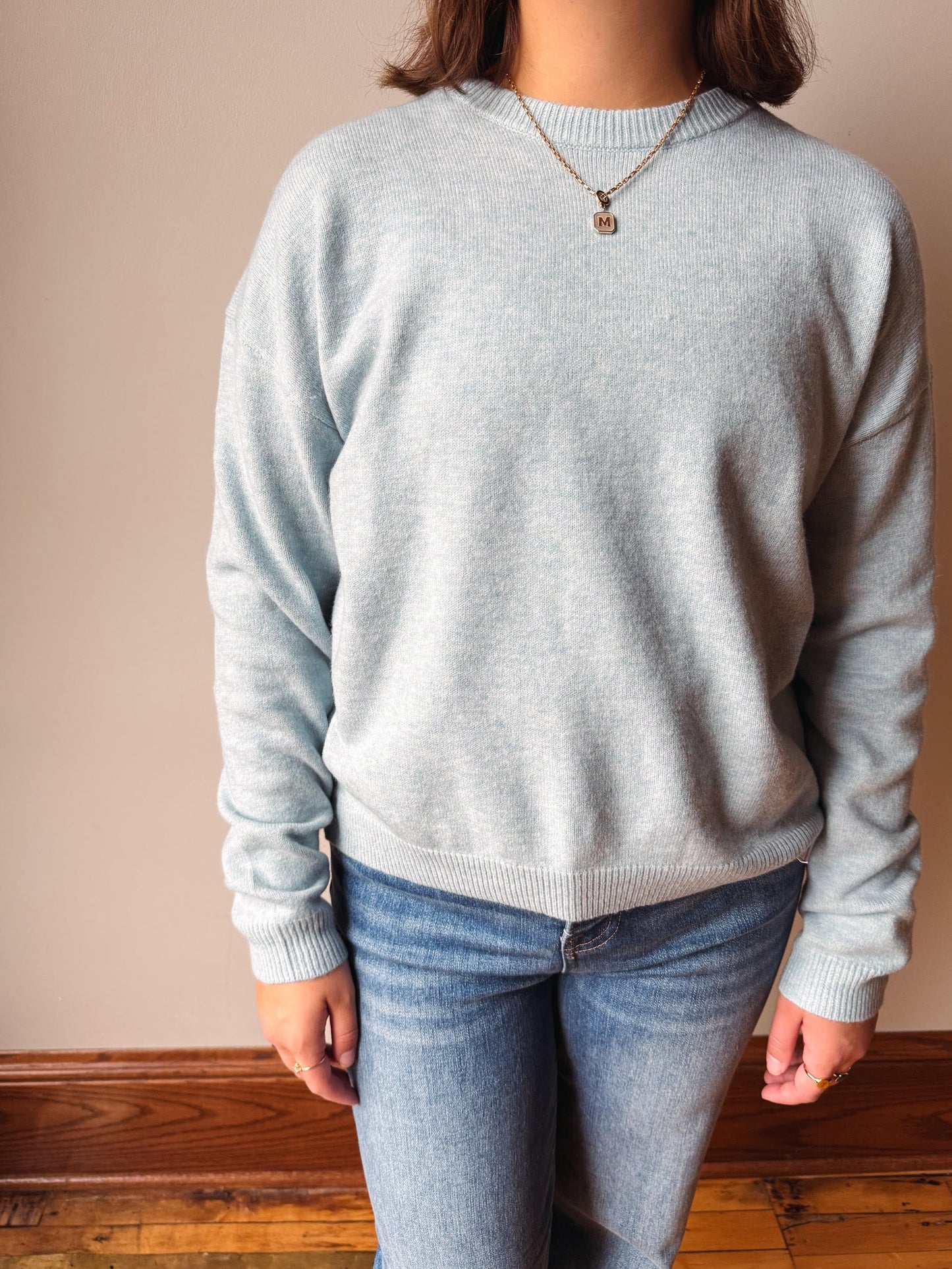 A person wearing a light heather gray crewneck sweater and blue jeans, standing against a beige wall with a wooden baseboard.
