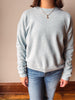 A person wearing a light heather gray crewneck sweater and blue jeans, standing against a beige wall with a wooden baseboard.