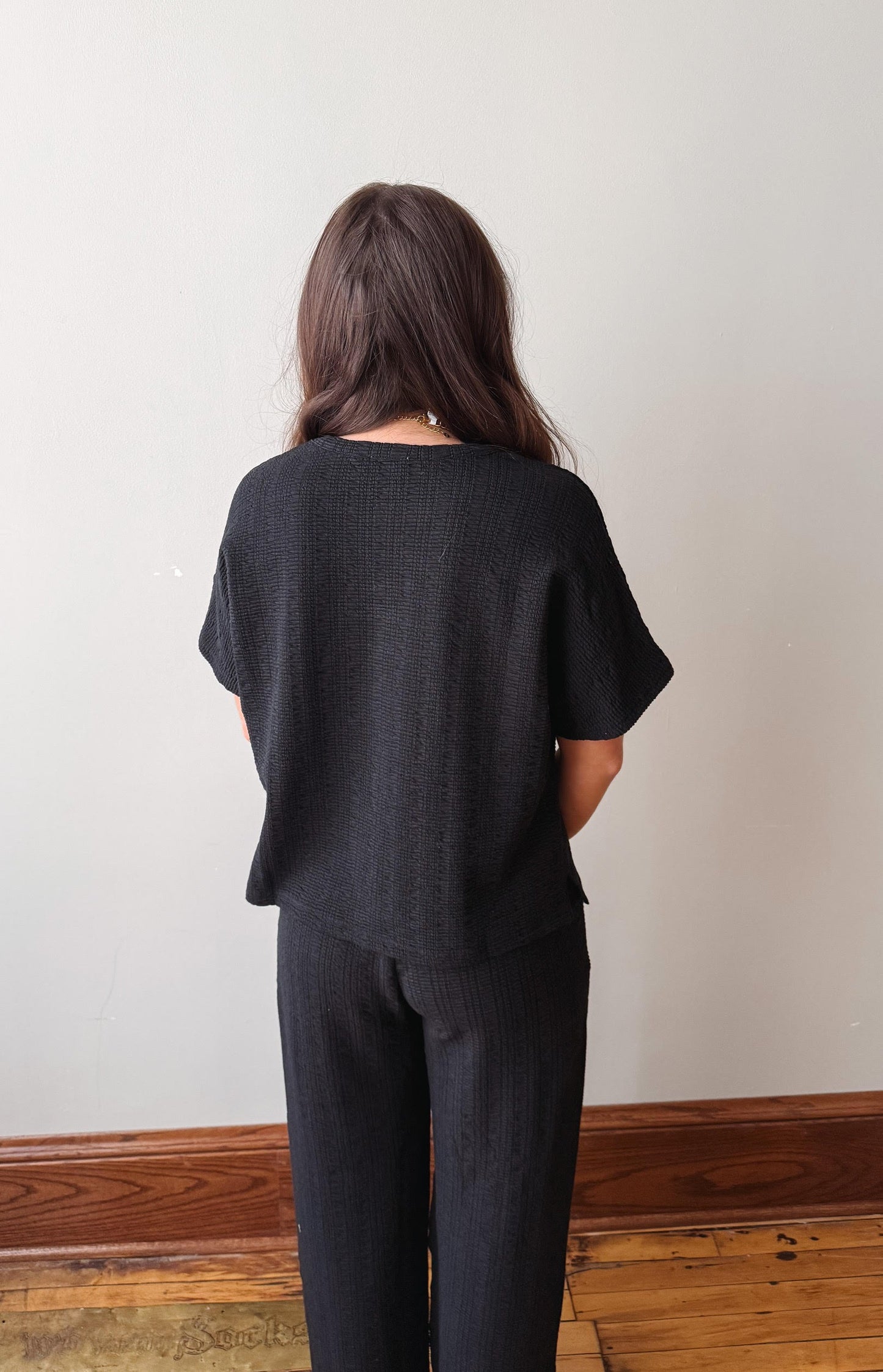Person wearing a black outfit standing against a plain white wall.