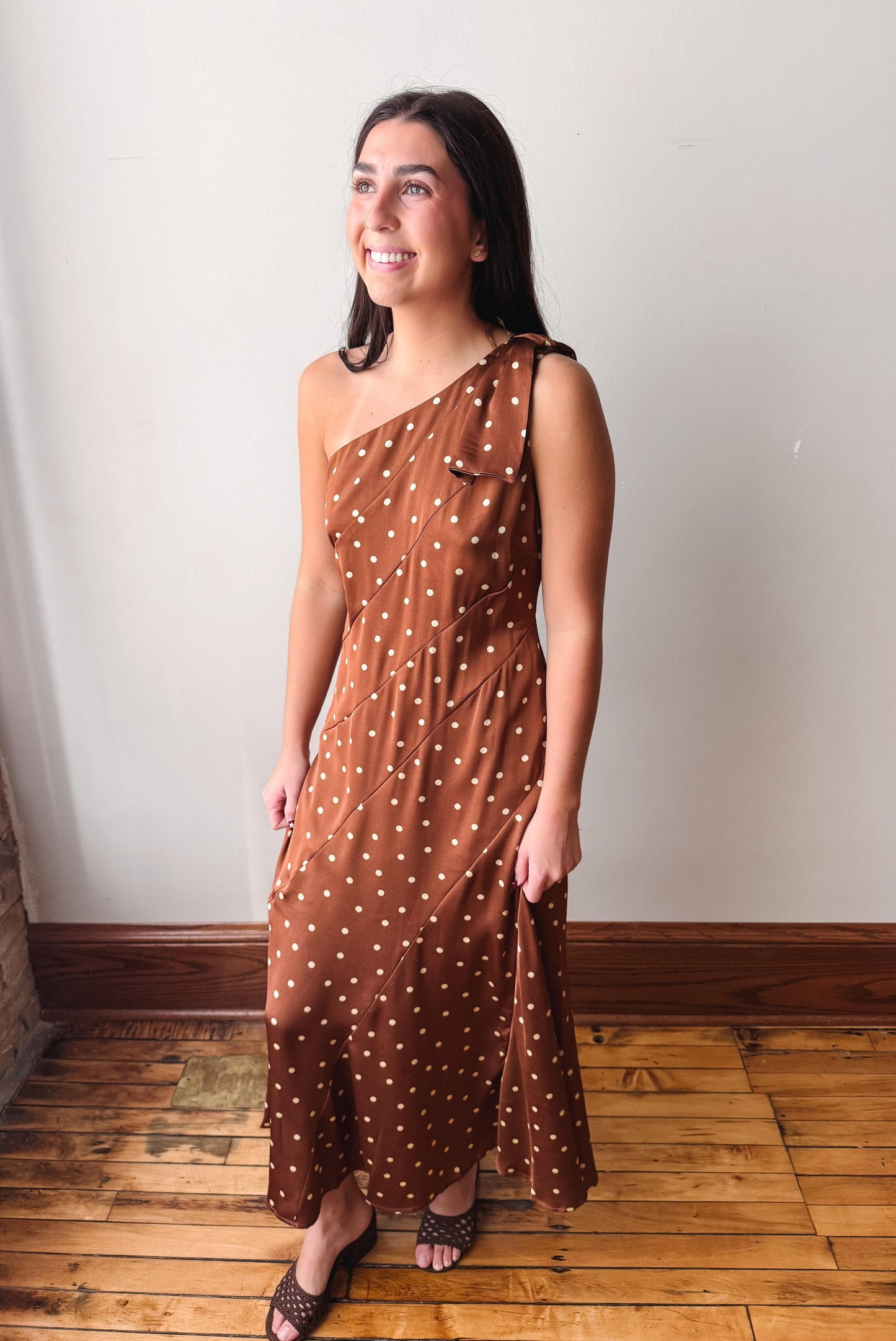 A woman modeling a brown one-shoulder midi dress with white polka dots, standing on a wooden floor against a plain off-white wall.