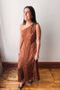 A woman modeling a brown one-shoulder midi dress with white polka dots, standing on a wooden floor against a plain off-white wall.
