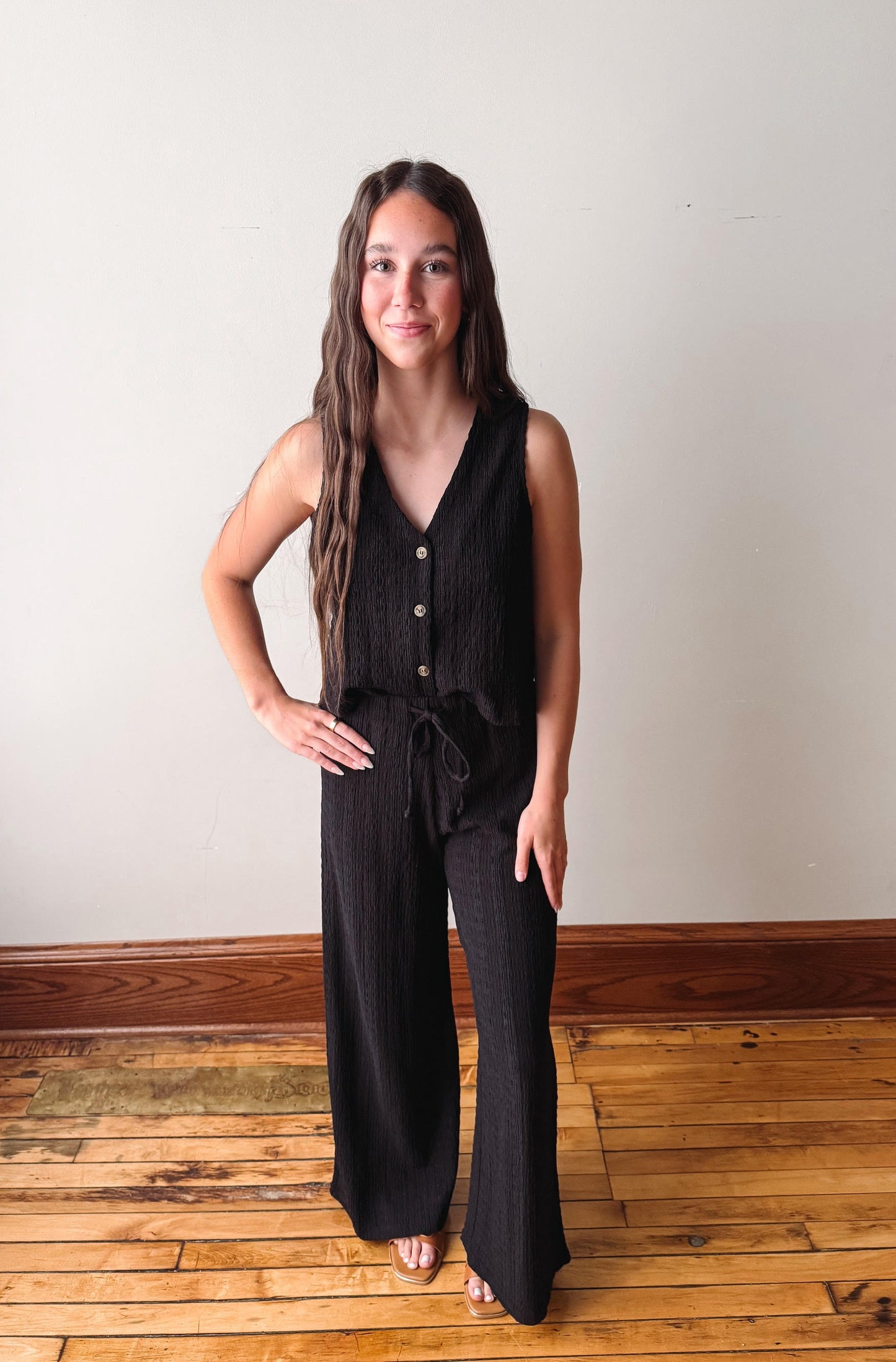 Woman wearing a black sleeveless jumpsuit standing on a wooden floor with a white wall background