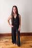 Woman wearing a black sleeveless jumpsuit standing on a wooden floor with a white wall background
