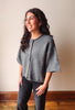 A woman with long dark wavy hair wearing a gray knit poncho-style cardigan and dark gray pants, standing in a room with a beige wall and wooden floor.