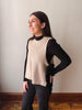 A woman modeling a sleeveless beige knit vest over a black long-sleeve mock neck top and black pants, standing against a plain light gray wall.