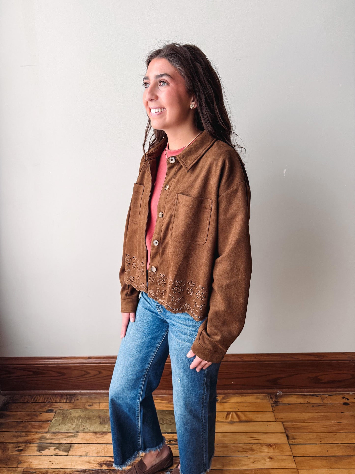 A woman wearing a brown suede-like button-up jacket over a coral-pink top and blue jeans, standing in a room with a light gray wall and wooden floor.