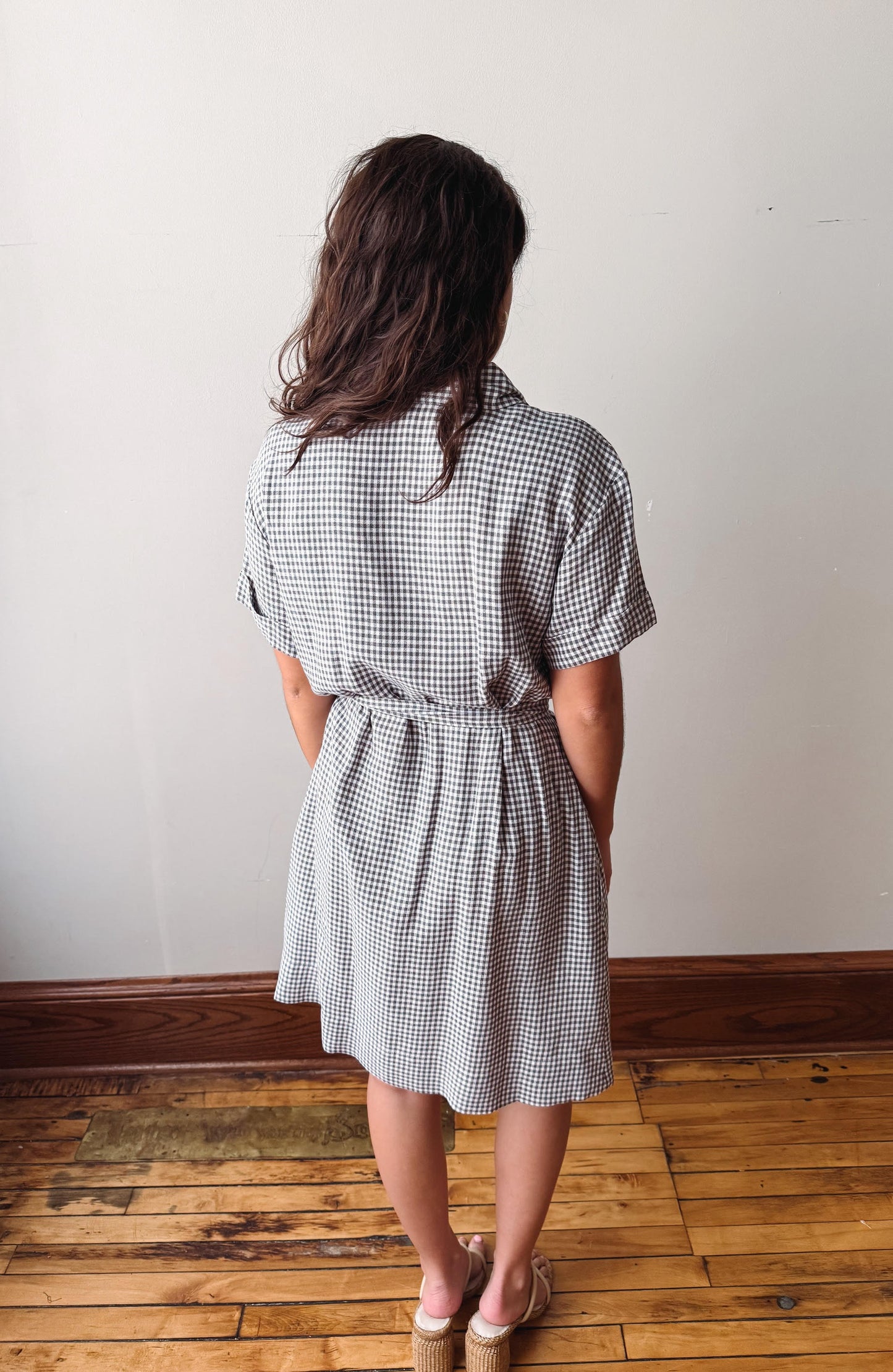 Woman wearing a checkered dress standing on a wooden floor with a white wall background