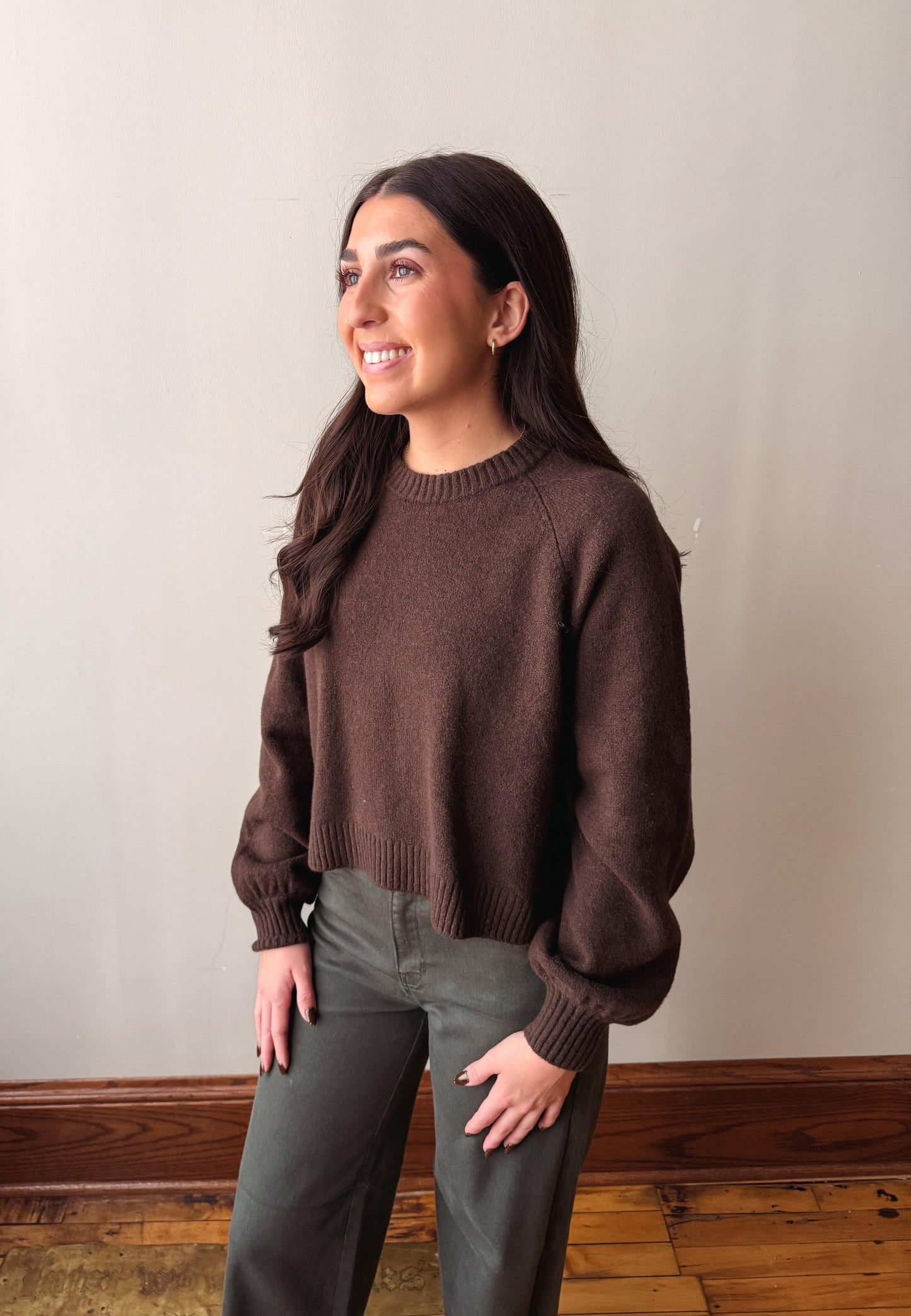 A woman wearing a dark brown knit crewneck sweater and olive green pants, standing in a room with a beige wall and wooden floor.