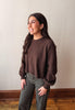 A woman wearing a dark brown knit crewneck sweater and olive green pants, standing in a room with a beige wall and wooden floor.