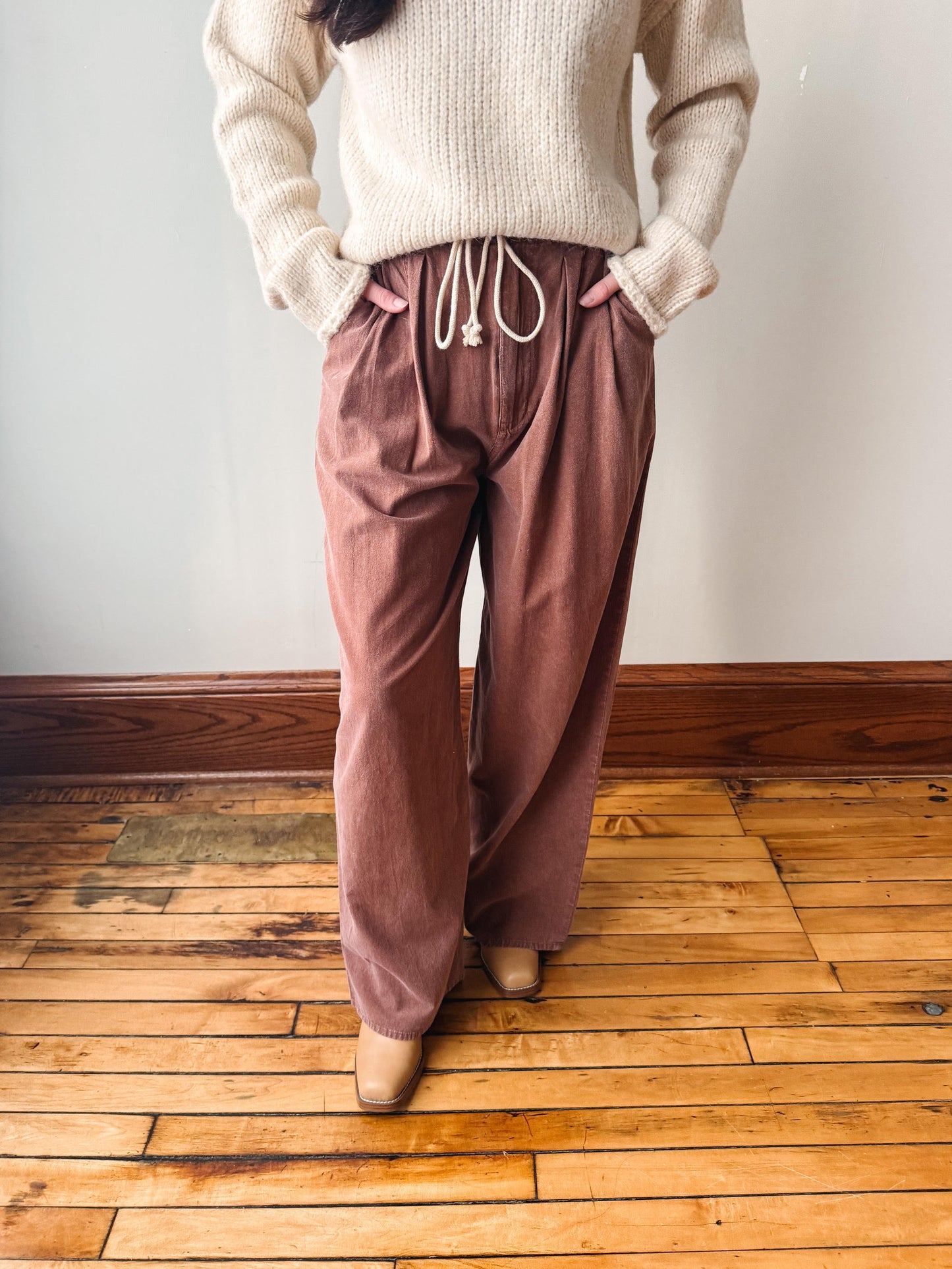 A person wearing loose, wide-leg dusty rose drawstring pants with a cream ribbed knit sweater, standing on a wooden floor against a plain wall.