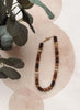 A beaded necklace with dark brown, medium brown, and cream beads, styled on a light pink textured fabric with eucalyptus leaves in the corner.