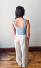 Person wearing a light blue tank top and beige pants standing on a wooden floor with a white wall background