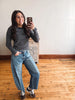 A woman takes a mirror selfie wearing a black and gray striped long-sleeve shirt, wide-leg blue jeans, and brown sneakers with white laces, standing in a room with a light-colored wall and wooden floor.