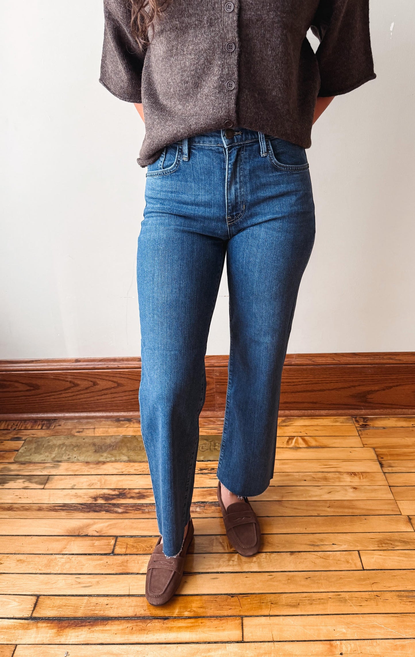 A person wearing medium-wash blue straight-leg jeans, a brown knit top, and brown suede loafers, standing on a wooden floor against a white wall.