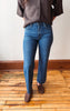 A person wearing medium-wash blue straight-leg jeans, a brown knit top, and brown suede loafers, standing on a wooden floor against a white wall.