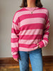 A person wearing a pink and light pink horizontally striped knit sweater with blue jeans, standing indoors against a plain wall.