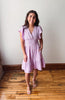 Woman wearing a light purple dress standing on a wooden floor with a white wall background