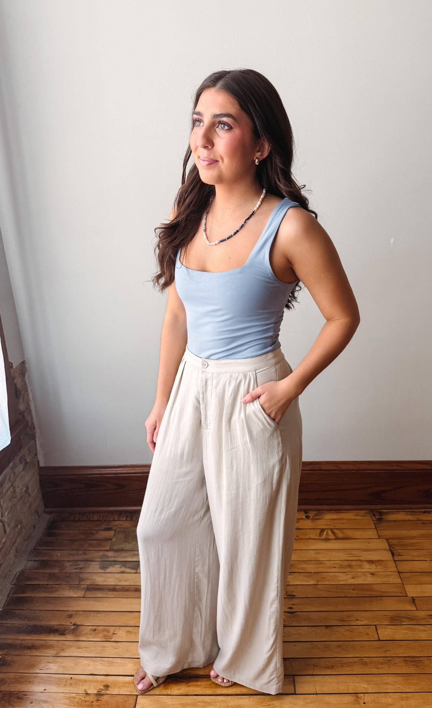 Woman wearing a light blue tank top and beige wide-leg pants standing in a room with wooden flooring.