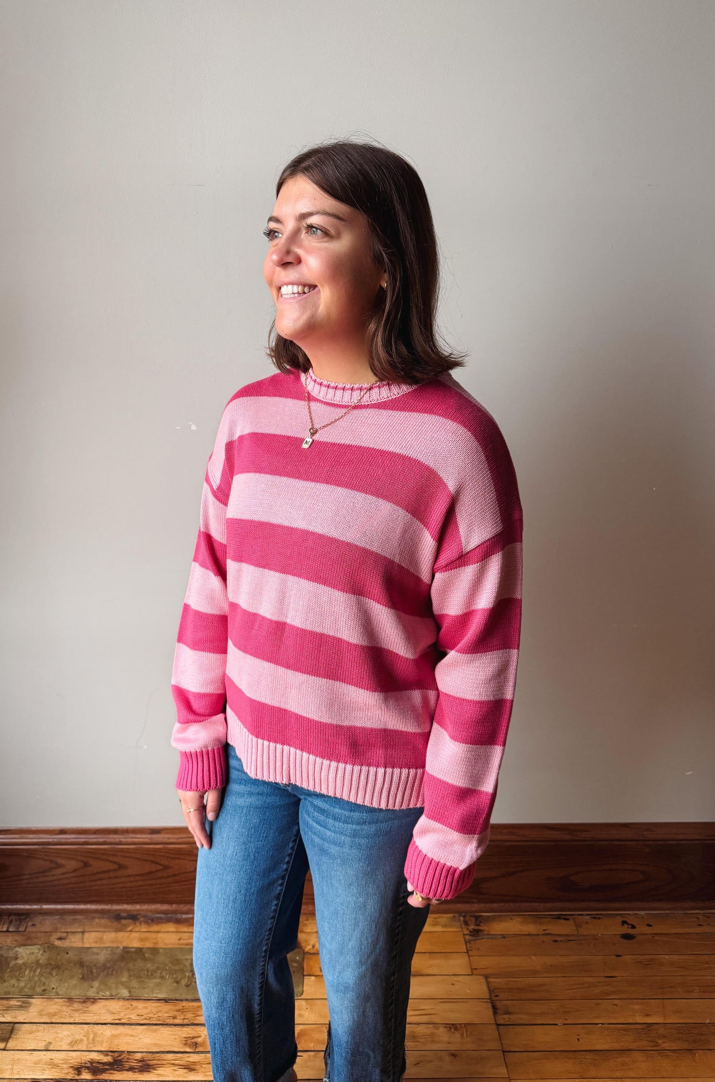 A woman wearing a pink and magenta striped knit sweater with blue jeans, standing in a room with a beige wall and wooden floor.
