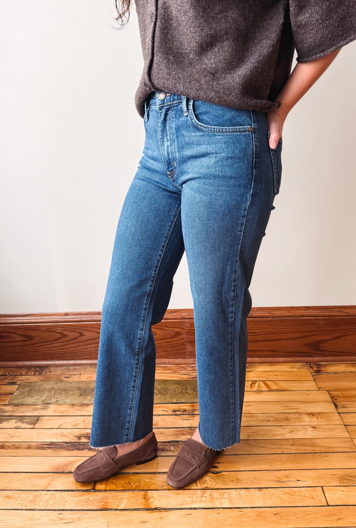 A person wearing blue straight-leg jeans, a dark gray knit top, and brown suede loafers, standing on a wooden floor against a plain off-white wall.