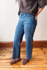 A person wearing blue straight-leg jeans, a dark gray knit top, and brown suede loafers, standing on a wooden floor against a plain off-white wall.
