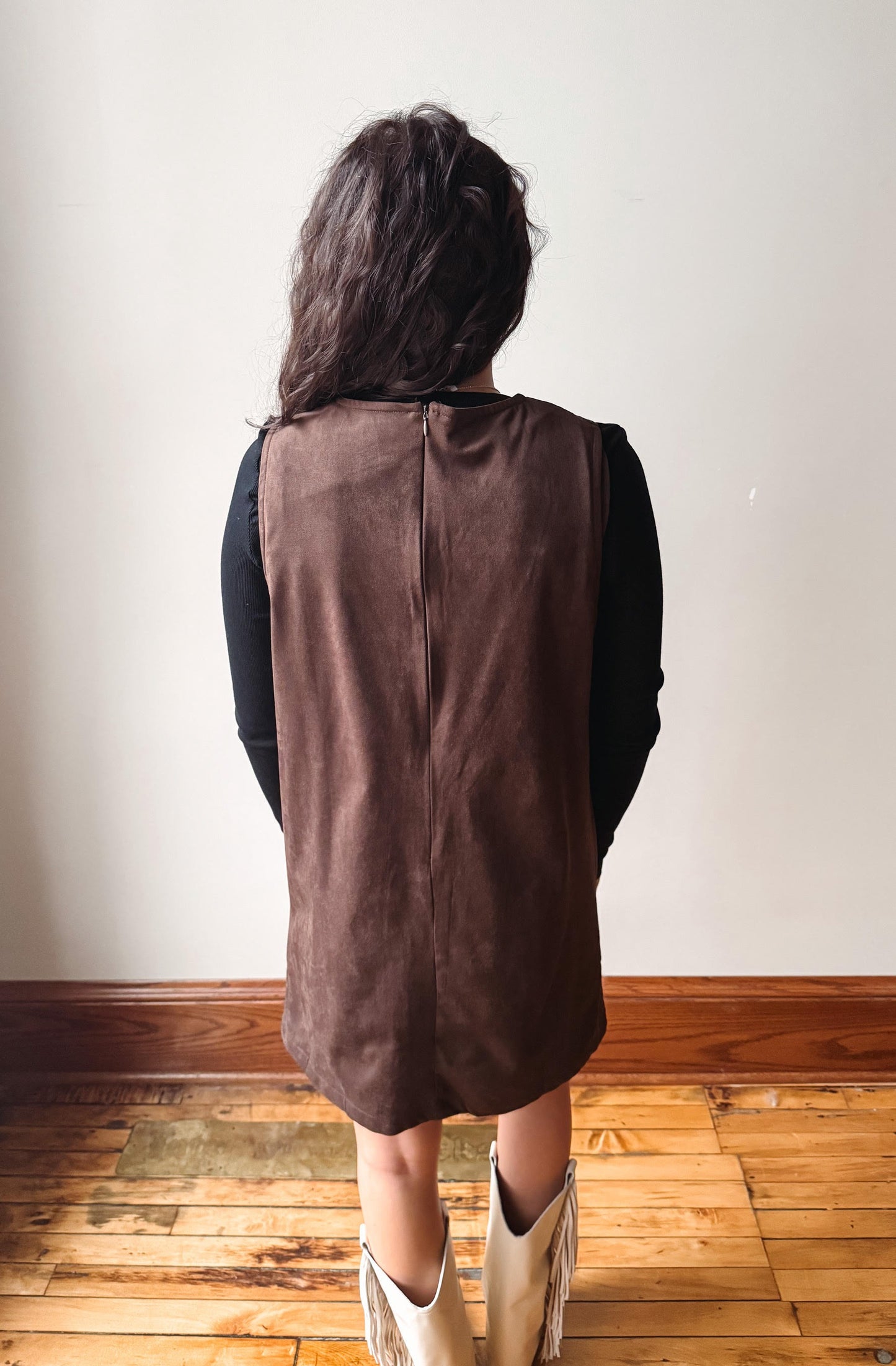 A person viewed from behind wearing a brown sleeveless vest over a black long-sleeve top, standing on a wooden floor against a plain off-white wall.