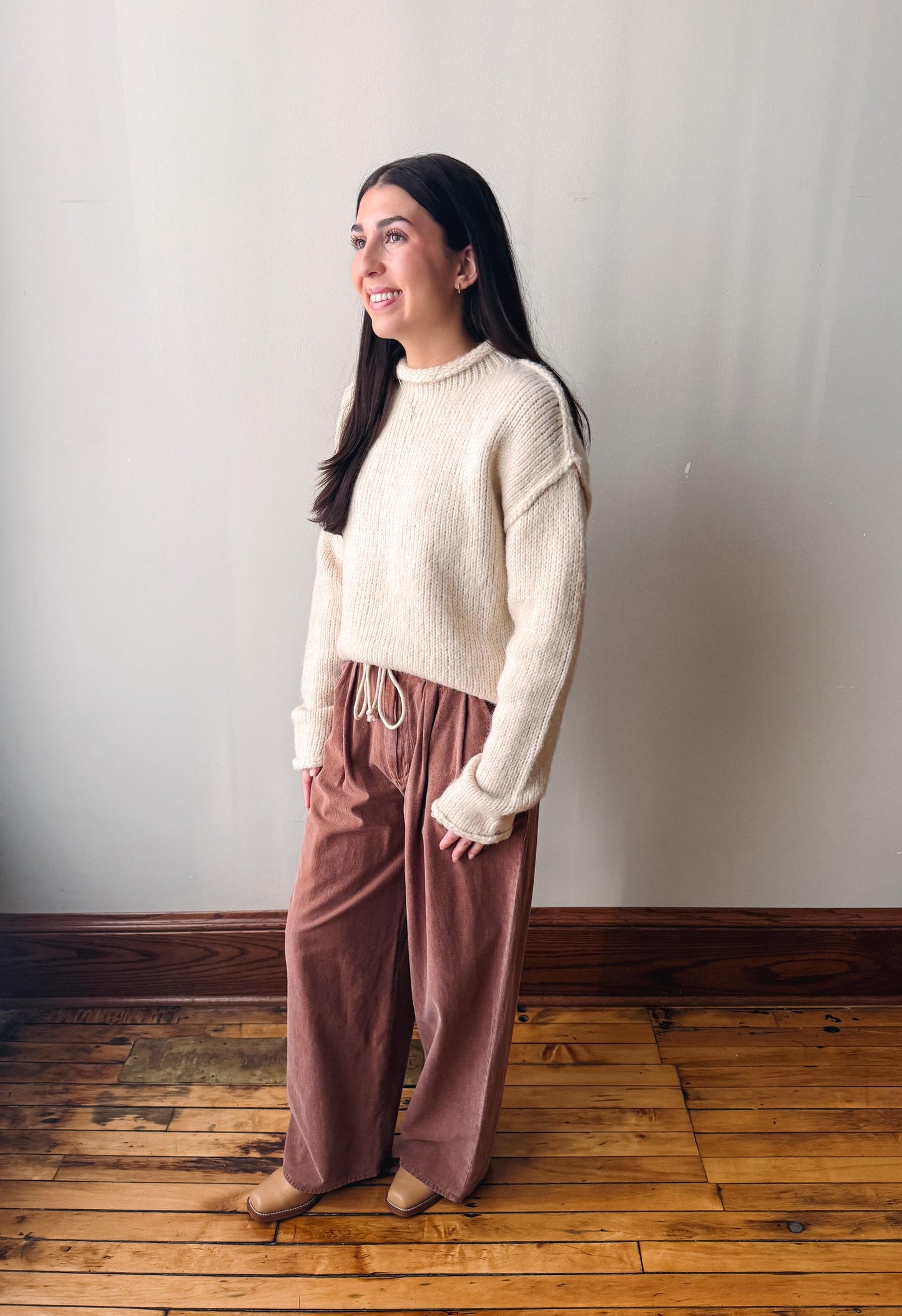 A woman stands in a room with a light gray wall and wooden floor, wearing a cream ribbed knit mock-neck sweater and dusty rose wide-leg drawstring pants.