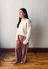 A woman stands in a room with a light gray wall and wooden floor, wearing a cream ribbed knit mock-neck sweater and dusty rose wide-leg drawstring pants.