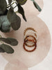 Three beaded stretch bracelets stacked together on a light beige fabric background with a watercolor circle and green eucalyptus leaves. The bracelets include white pearl-like beads, gold-toned metallic beads, and brown translucent bamboo-style rings with gold-toned connectors.