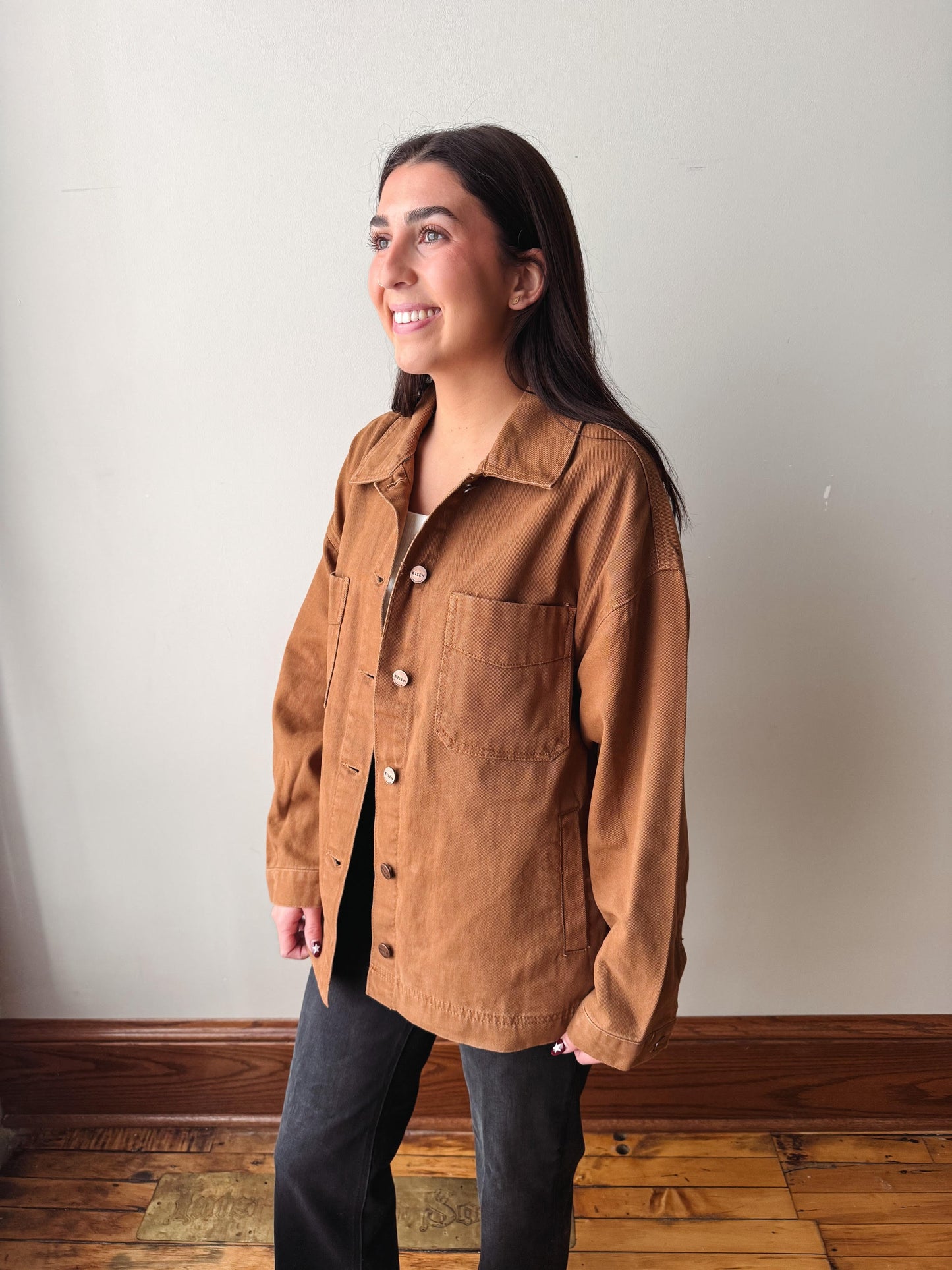 A woman wearing a brown corduroy button-up shirt jacket and black pants, standing in a room with a light gray wall and wooden floor.
