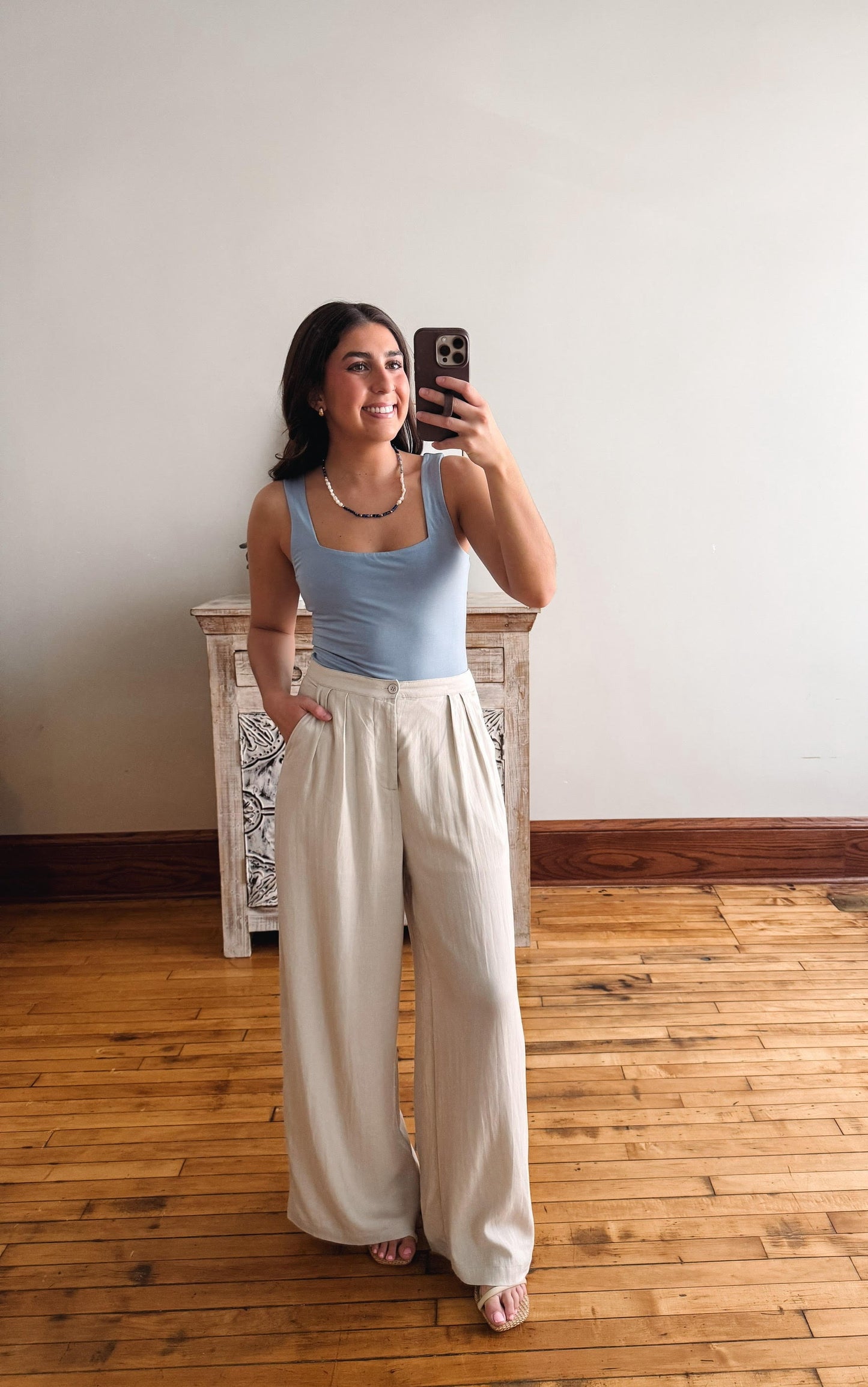 Woman taking a mirror selfie wearing a light blue top and beige wide-leg pants in a room with wooden flooring.