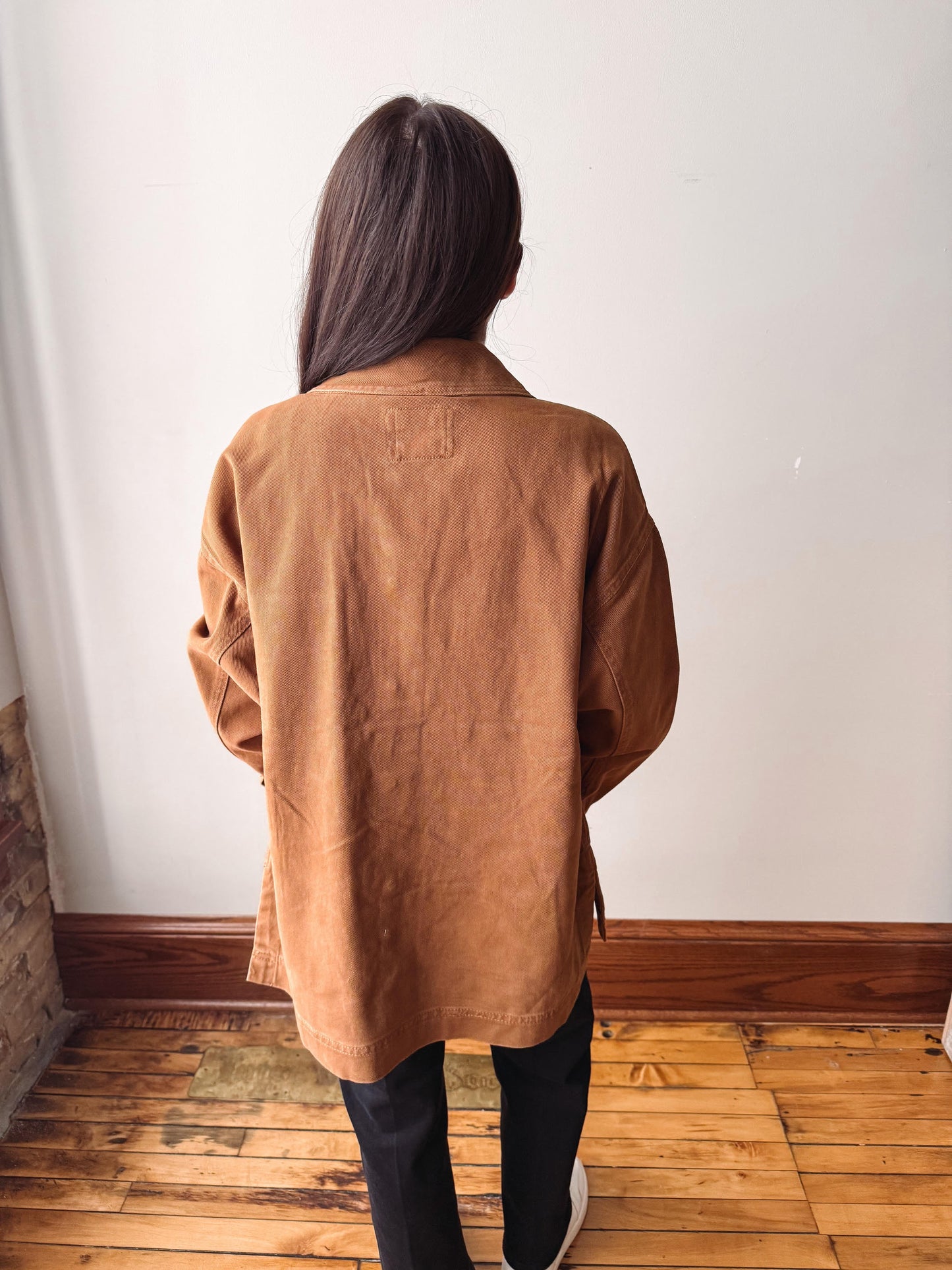 A person viewed from behind wearing an oversized tan-brown linen jacket, black pants, and white sneakers, standing on a wooden floor against a white wall.