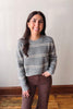 A woman smiling and wearing a gray knit sweater with horizontal cream stripes and brown pants, standing against a plain off-white wall with a wooden baseboard and wood floor.