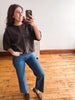 A woman takes a mirror selfie indoors wearing a dark brown textured short-sleeve top, blue jeans, and brown loafers, standing on a wooden floor against a plain wall.