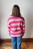 A person viewed from behind wearing a pink and light pink striped knit sweater and blue jeans, standing against a plain off-white wall with a wooden floor visible.