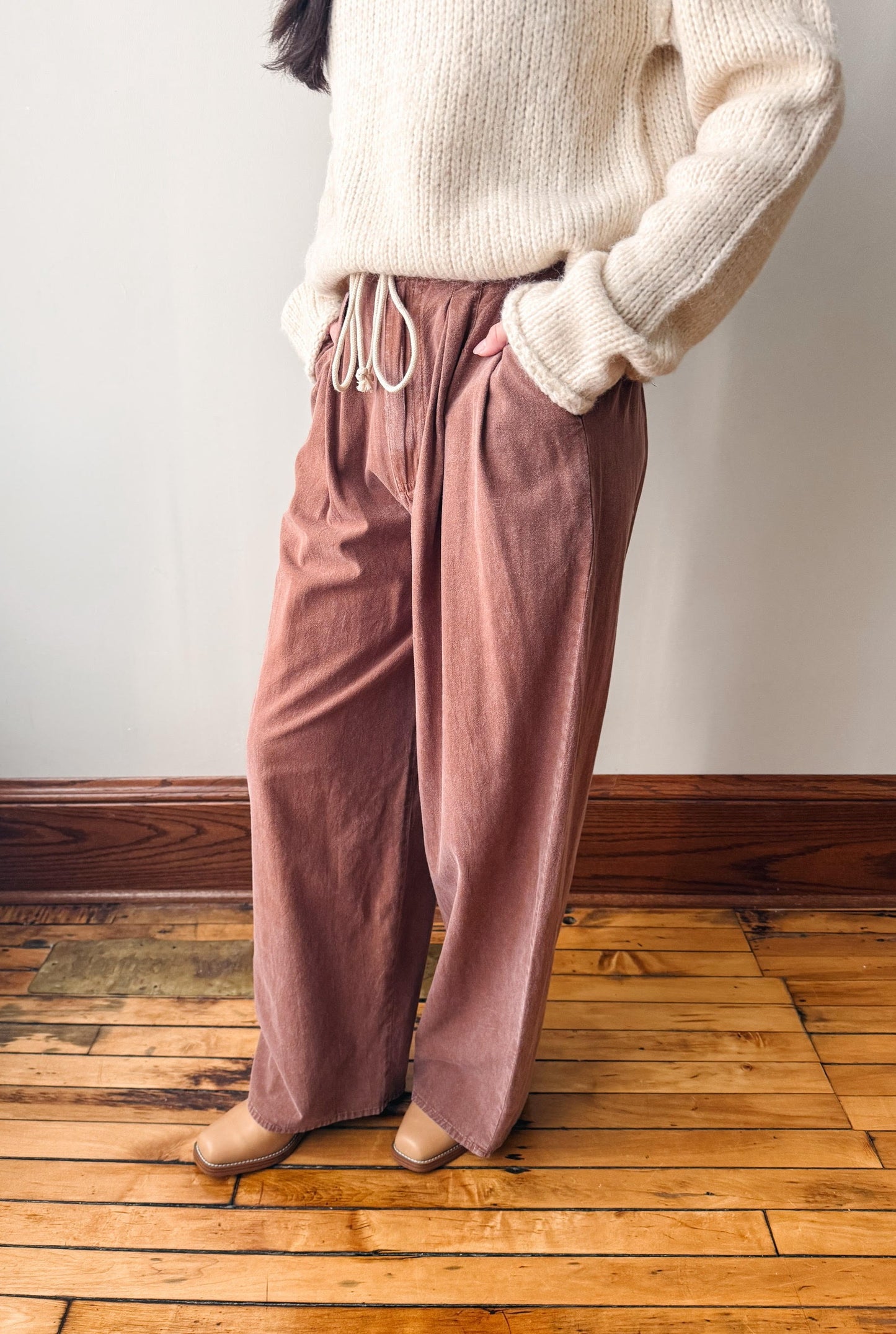 A person wearing dusty rose wide-leg drawstring pants with a cream knit sweater, standing on a wooden floor against a plain wall.