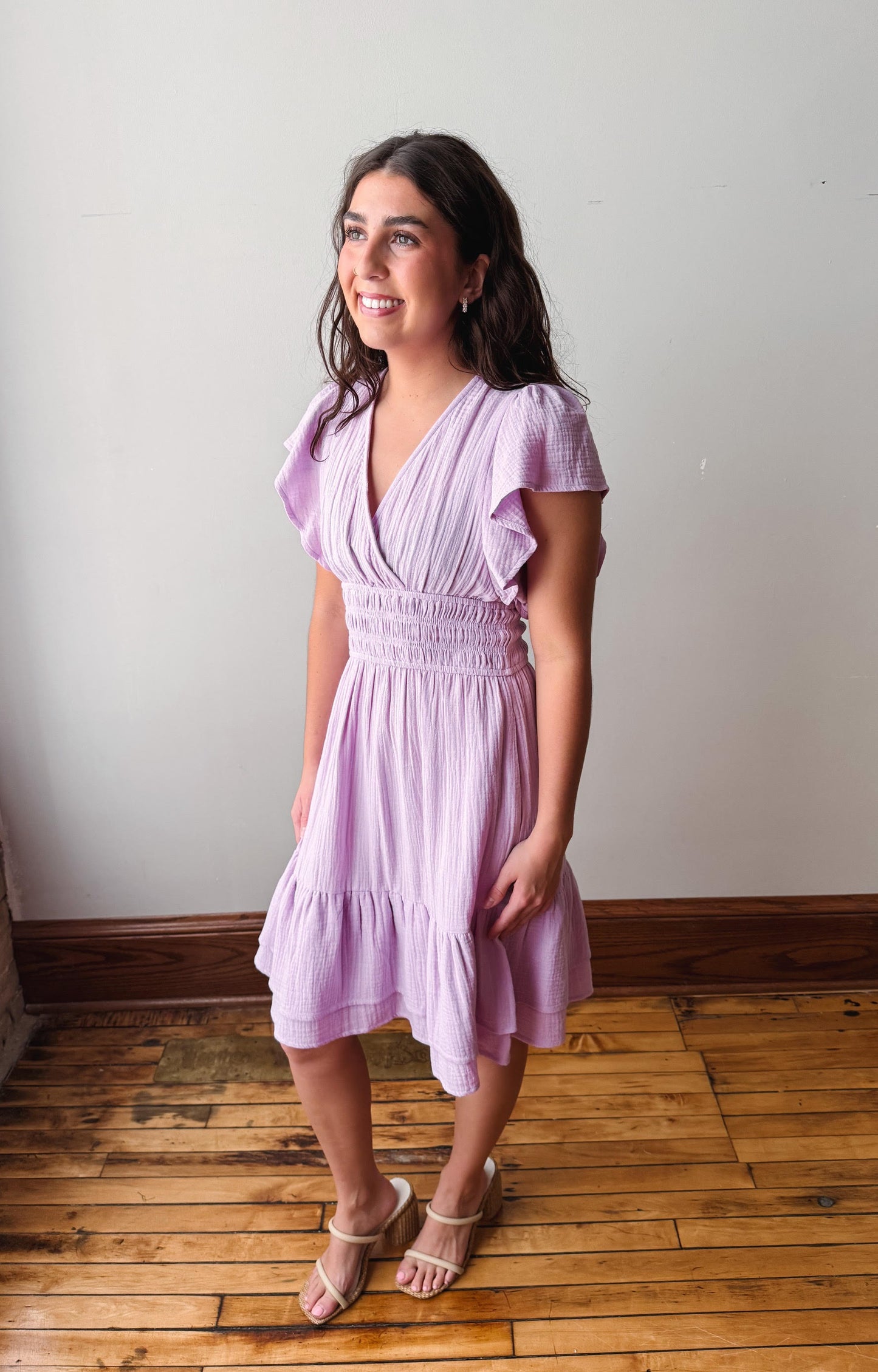 Woman wearing a light purple dress standing on a wooden floor against a white wall.