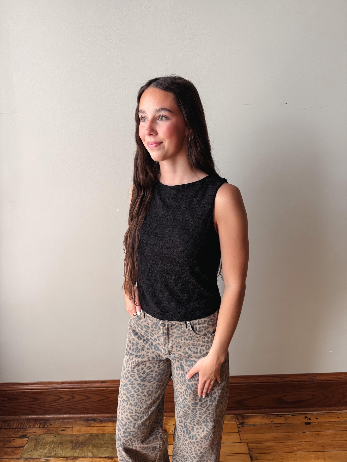 Woman wearing a black sleeveless top and patterned pants standing against a plain wall.